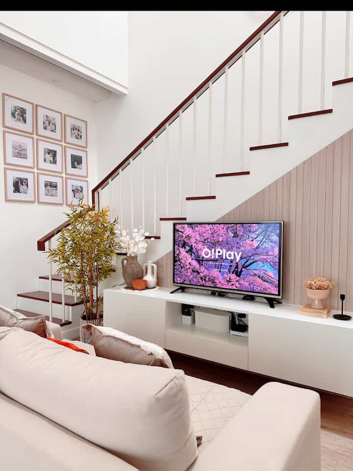 Bright white and beige toned living room featuring a beige sofa, TV stand, picture frames, and plants creating a simple and warm atmosphere