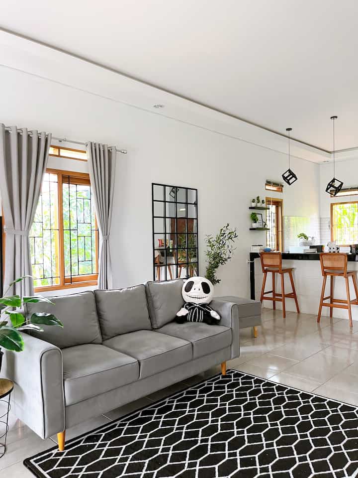 Simple living room with gray sofa and monotone carpet on white walls and tiled floor, featuring wooden bar stools and a black kitchen counter