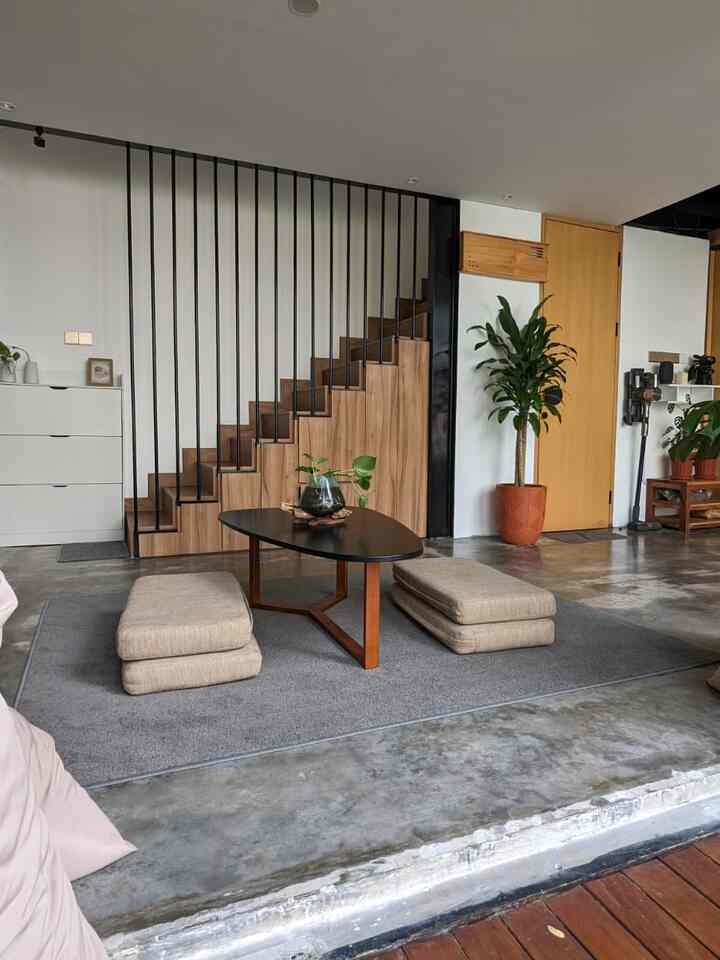 Modern natural studio apartment with white walls and brown wood stairs, featuring a black coffee table and cushions on a gray rug creating a simple, spacious atmosphere