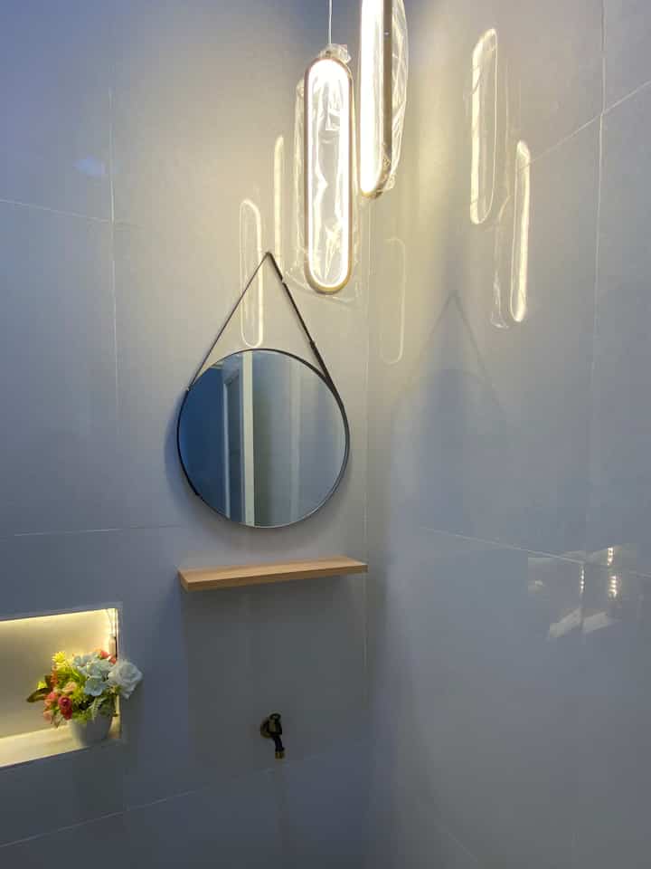 Minimal bathroom corner with white walls, wood-tone shelf, black round mirror, and pendant lights