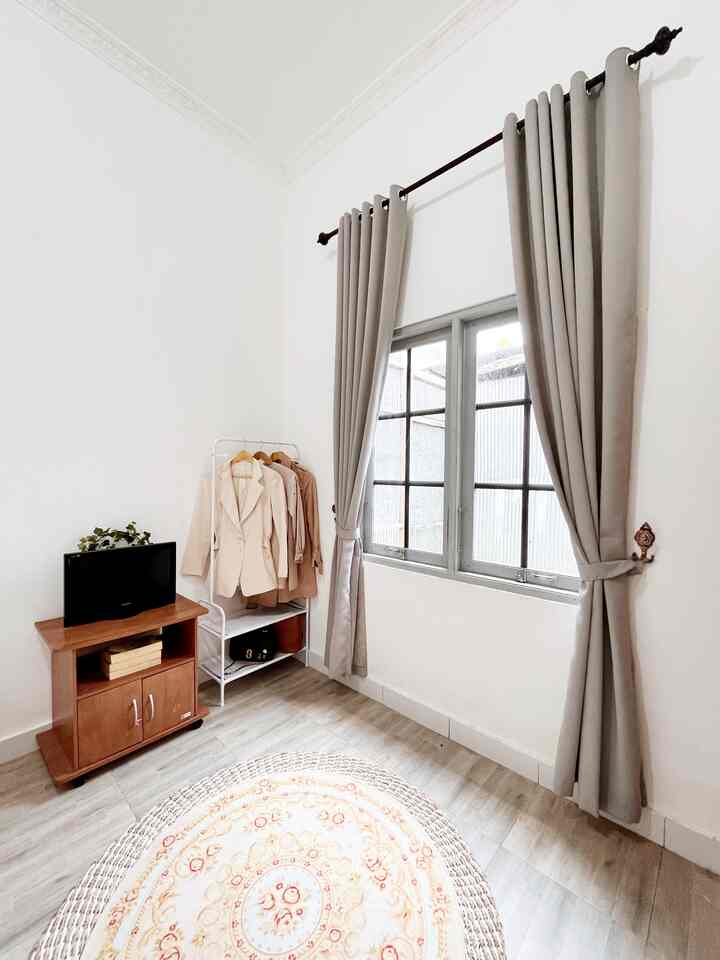 White and brown toned, long narrow small bedroom with curtains and clothes rack by window, creating cozy space