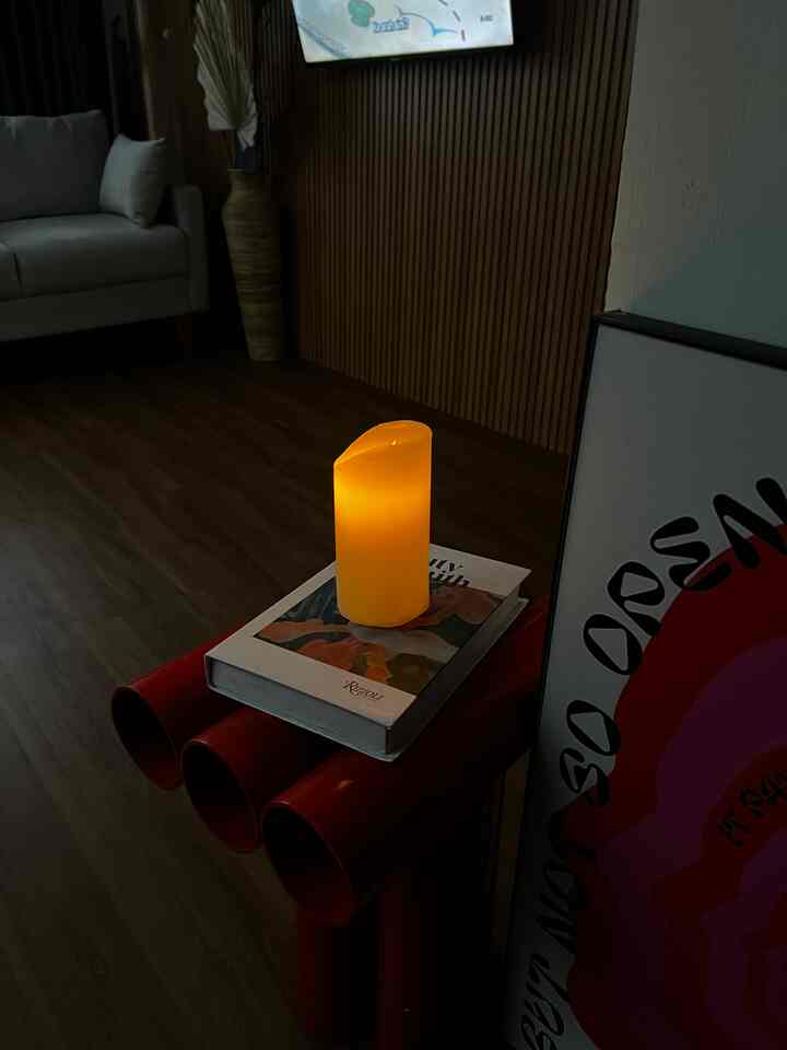 Living room with gray sofa, wooden slatted wall, red tubular side table topped with glowing yellow candle and a book in dim lighting