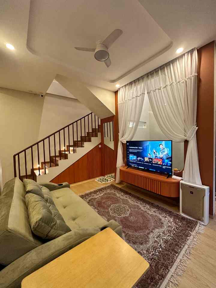 A natural-tone small living room featuring a green sofa, brown patterned carpet, white curtains, and staircase on the side
