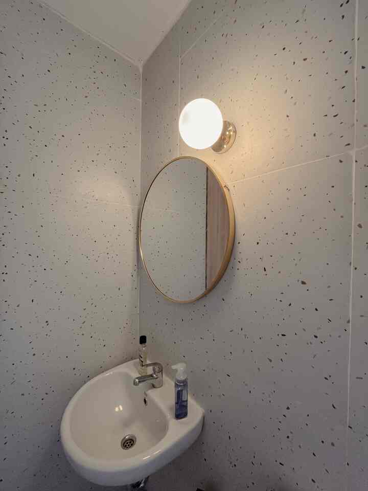 White terrazzo-tiled bathroom featuring a round gold-framed mirror and wall sconce, creating a clean atmosphere