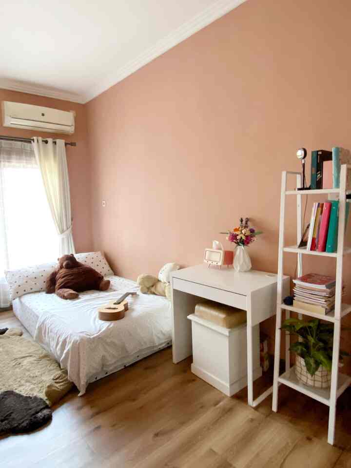 Dusty pink walled, wood-tone floored long narrow kids' room featuring a floor mattress bed and white desk, creating a cozy atmosphere
