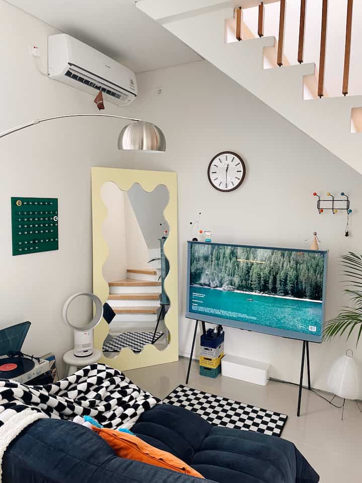 Monotone living room featuring a black two-seater sofa, large wavy mirror, TV, and clock in a modern setting