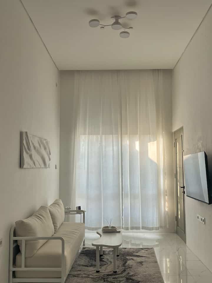 White and beige toned living room featuring a simple sofa, long coffee table, and bright natural light through sheer curtains