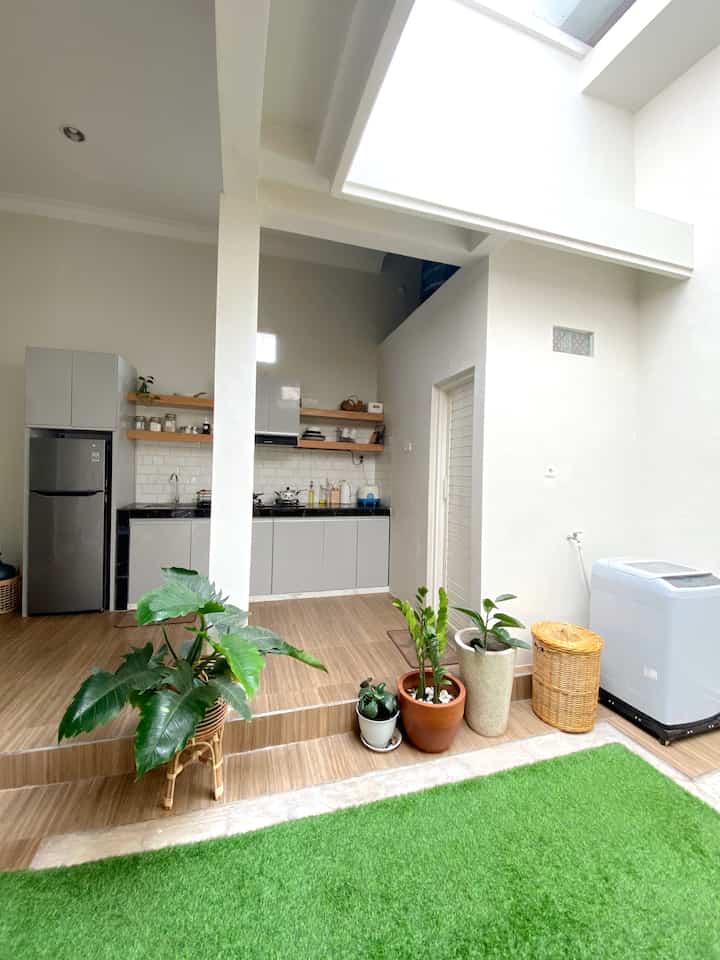 Bright and clean kitchen and laundry space in white and brown tones, featuring synthetic grass carpet and multiple potted plants arranged harmoniously