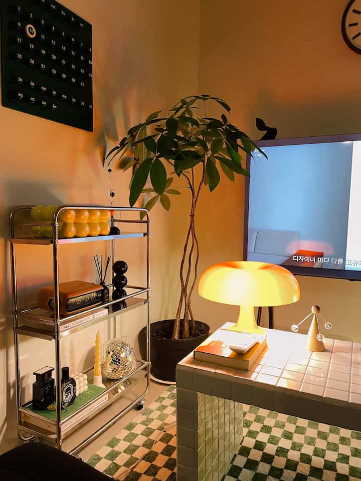 Cozy retro living room featuring warm orange lighting, green checkered carpet, a bubble-shaped table lamp, and a large indoor plant