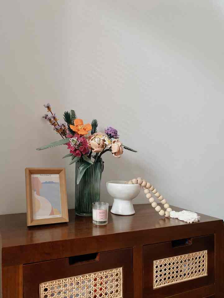 A natural wood-toned table corner featuring a vase of colorful flowers, candle, and framed artwork creating a simple cozy nook