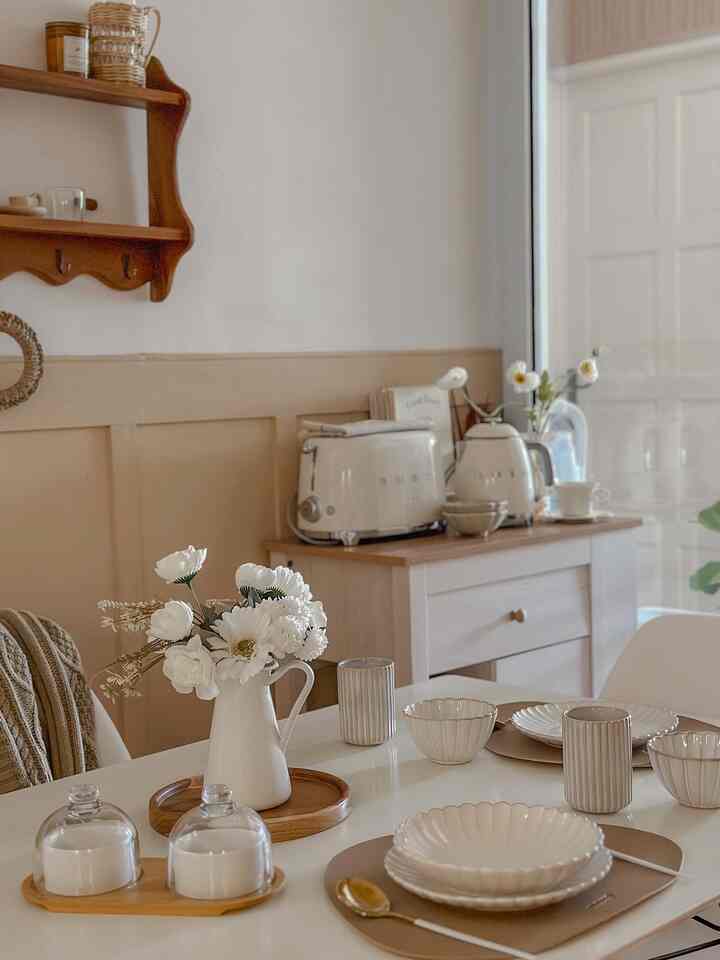 White and beige toned kitchen-dining space featuring simple white tableware and a vase on the dining table, creating a cozy atmosphere