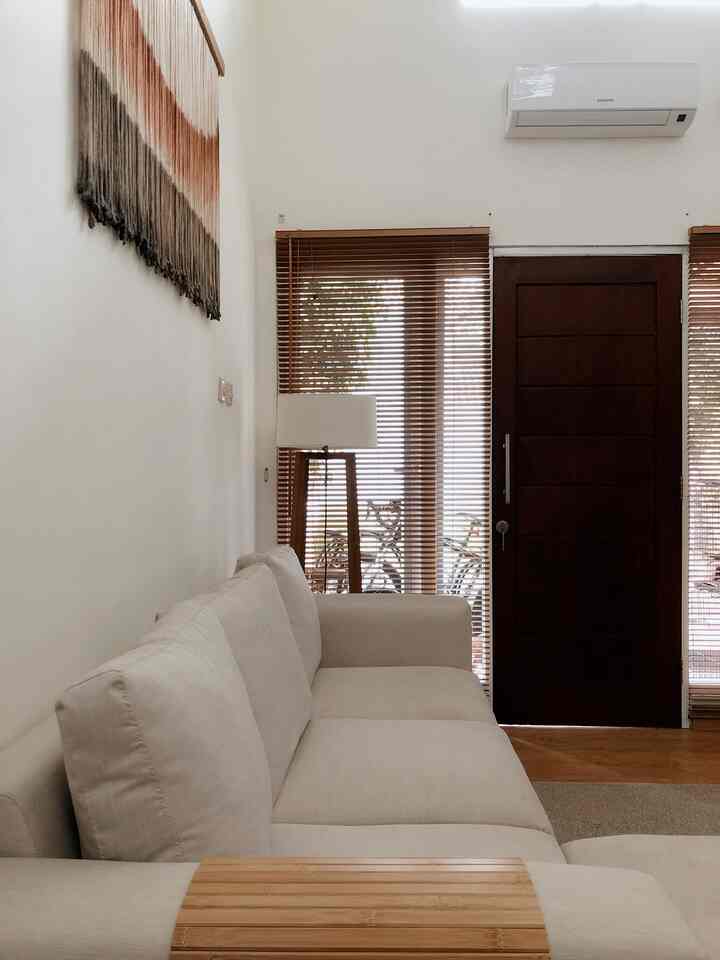 Natural tone living room featuring a beige sofa, brown wood-tone blinds, and a wooden entrance door with a simple interior