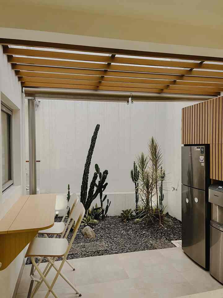 Bright semi-outdoor kitchen featuring natural wood-toned ceiling beams, stainless steel refrigerator, and a small bar table with two folding chairs, clean and minimalist atmosphere