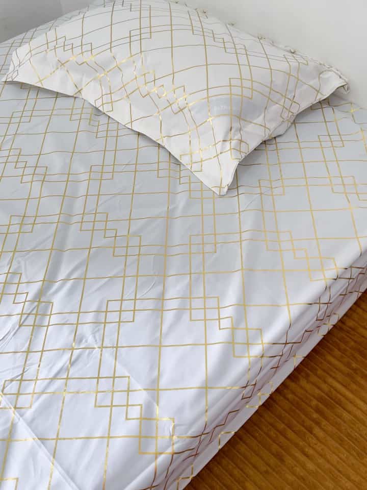 White and gold grid patterned bedding on bed in small bedroom featuring wooden floor and cabinet for cozy atmosphere