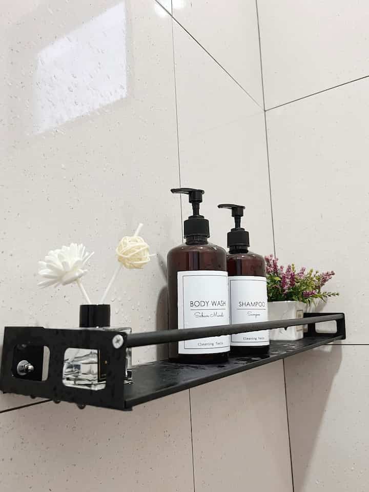 Beige tiled bathroom wall with black metal shelf holding shampoo, body wash bottles, and diffuser
