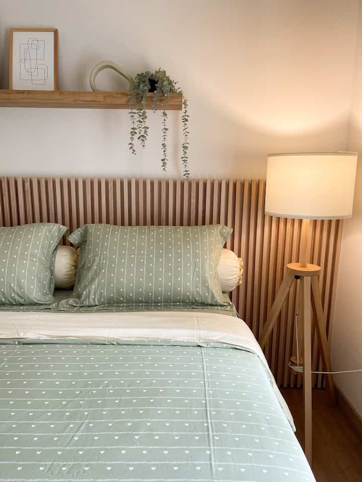 Natural wood tone bedroom with soft green bedding, featuring a wooden floor lamp and wall shelf with hanging plant, creating a cozy atmosphere