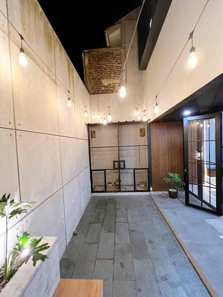 Gray concrete walls and flooring with black metal-framed glass doors create an industrial-style outdoor cafe space illuminated by hanging string lights