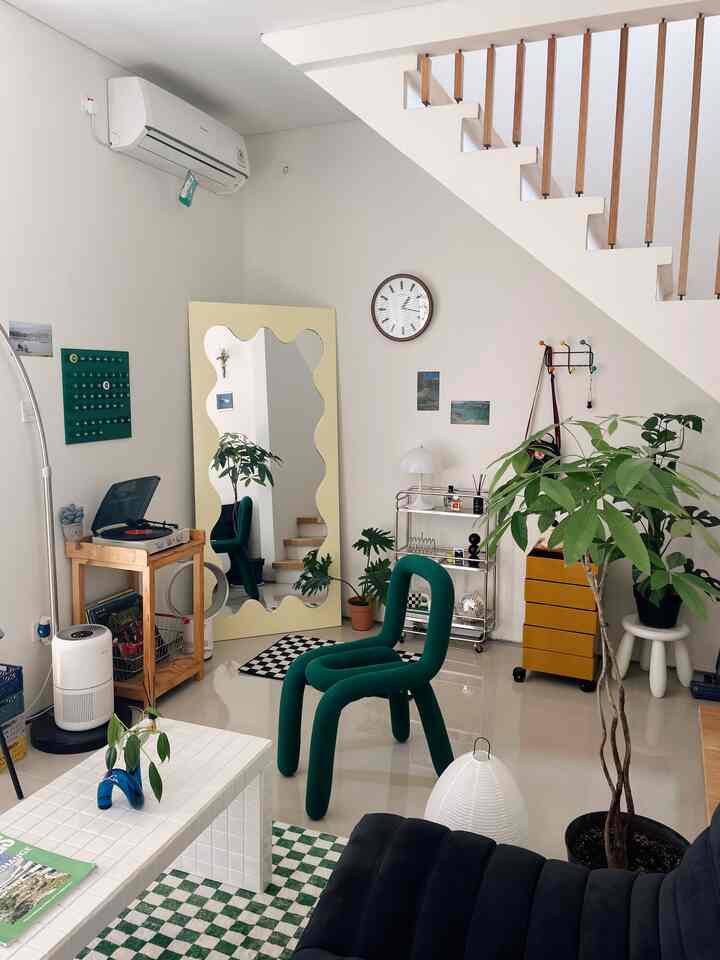 Bright white-toned living room featuring a unique green chair in center, large yellow wavy mirror on wall, plants and storage furniture arranged