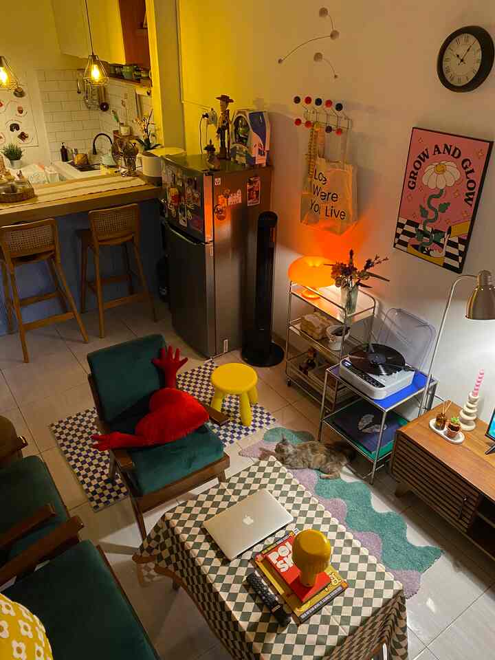 Cozy small living room and kitchen space featuring green mid-century modern furniture, colorful rugs, and warm yellow lighting