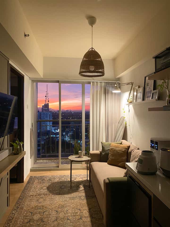 Natural beige-toned living room with a beige sofa, patterned rug, and balcony window showcasing a sunset view in a cozy atmosphere