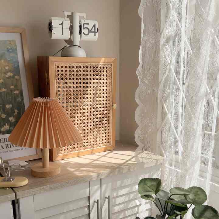 Bright white and natural wood-tone apartment corner featuring a flip clock, rattan storage cabinet, and pleated beige lamp creating a cozy atmosphere