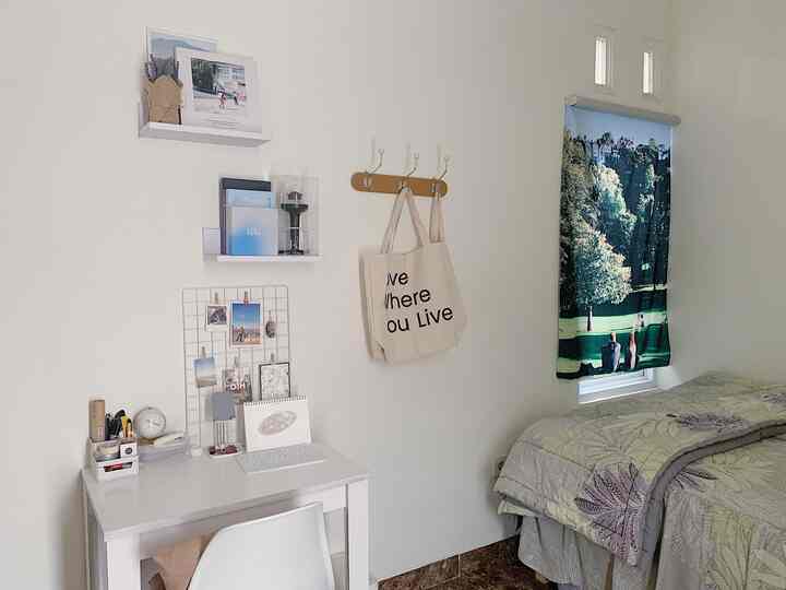 White-toned bedroom featuring a desk with wall decorations on left and a bed with scenic curtain on right, creating a simple space