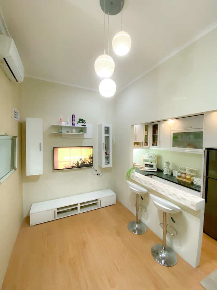 A white and wood-tone living room merged with kitchen featuring modern bar stools and pendant lights in a compact, minimalist space