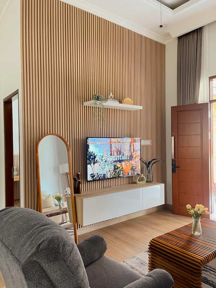 Wood tone and white living room featuring a wall-mounted TV and shelf center, full mirror left, gray armchair foreground; warm cozy vibe