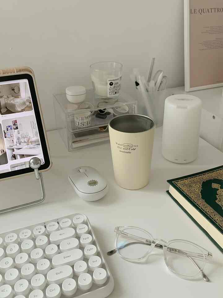 Minimal home office with white desk featuring white keyboard, mouse, beige tumbler, diffuser, glasses, and a book