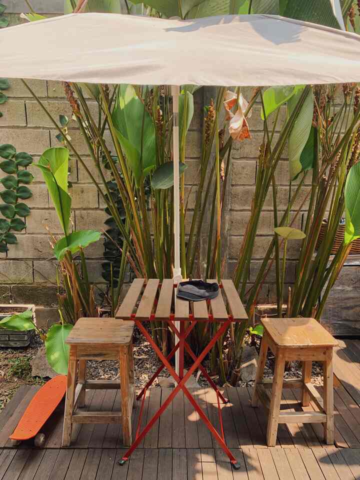 Natural-toned small outdoor deck with beige umbrella, wooden stools, and red metal-legged table creating a cozy backyard setting