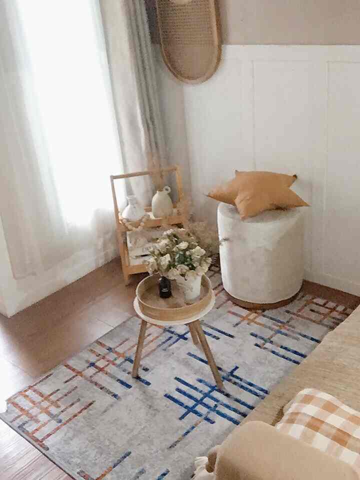 Natural-toned living room featuring a patterned rug centrally placed, round stool with beige cushion, soft curtains creating a cozy atmosphere