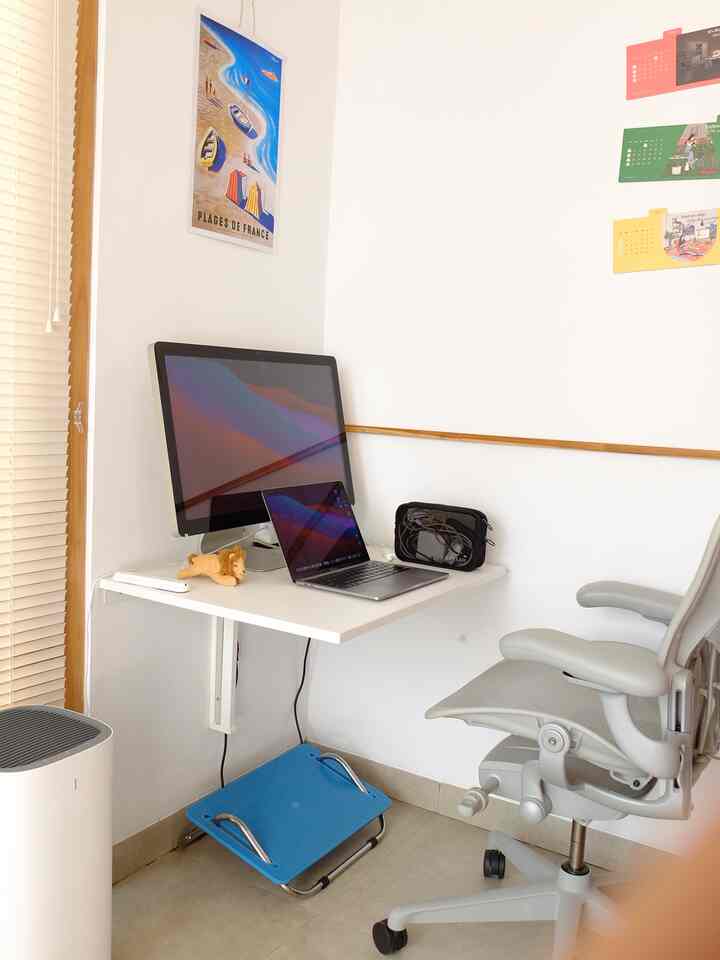 White-toned compact home office space featuring a wall-mounted desk with laptop and monitor, and a gray office chair creating a clean work environment