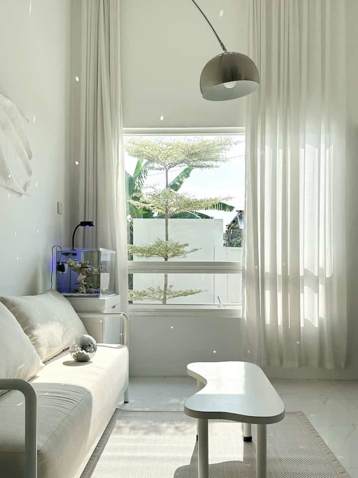 Bright white-toned living room featuring a minimalist sofa, unique coffee table, large window, and floor lamp in a clean space