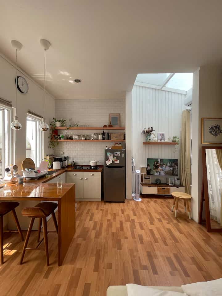 A white-walled studio apartment with wood-tone floors, featuring a minimalist kitchen and living area with stools, creating a cozy and spacious feel