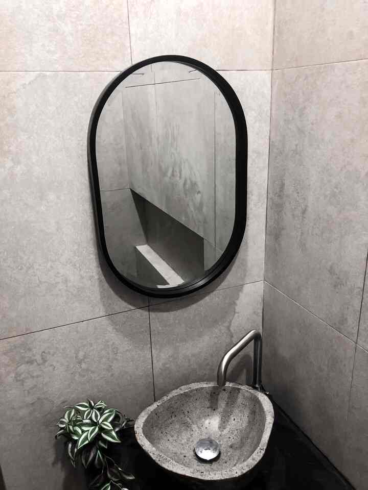 Natural tone bathroom featuring gray stone tiled walls, a stone sink, an oval black-framed mirror, and a small potted plant with a modern feel
