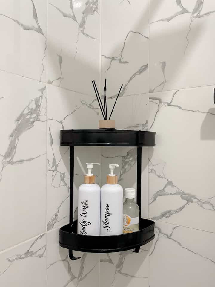 Simple bathroom space featuring black corner shelf with diffuser and shampoo bottles against white marble-patterned tiled wall