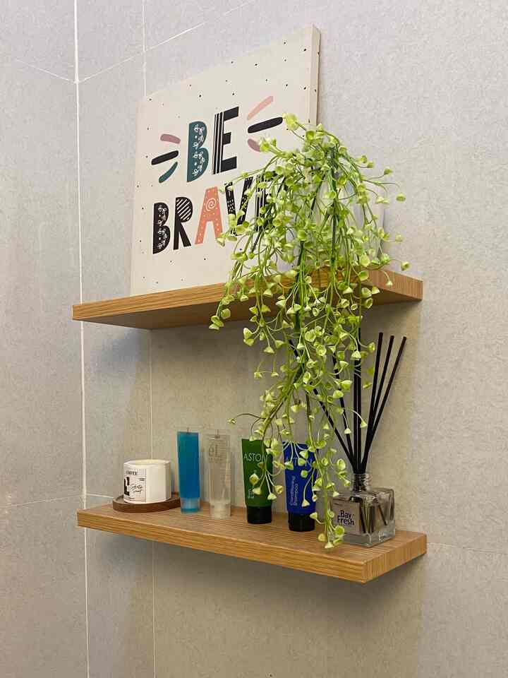 Bathroom corner with light gray tiled walls featuring wooden floating shelves holding plants and toiletries, creating a natural modern vibe