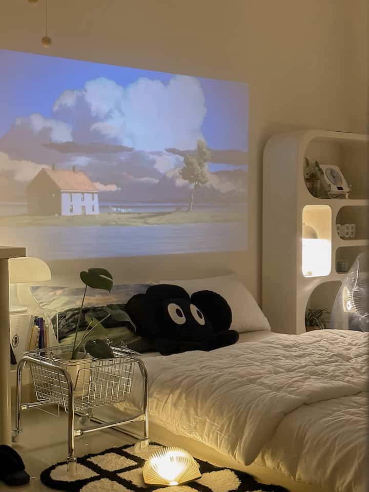White and beige toned bedroom with central bed featuring navy cushions, warm lighting, and a projected image on the wall creating cozy ambiance