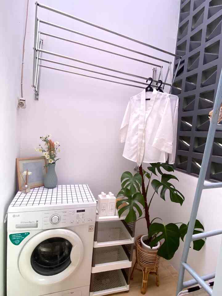 White-toned square small laundry space featuring a washing machine, shelving, and a large leafy plant in a simple arrangement