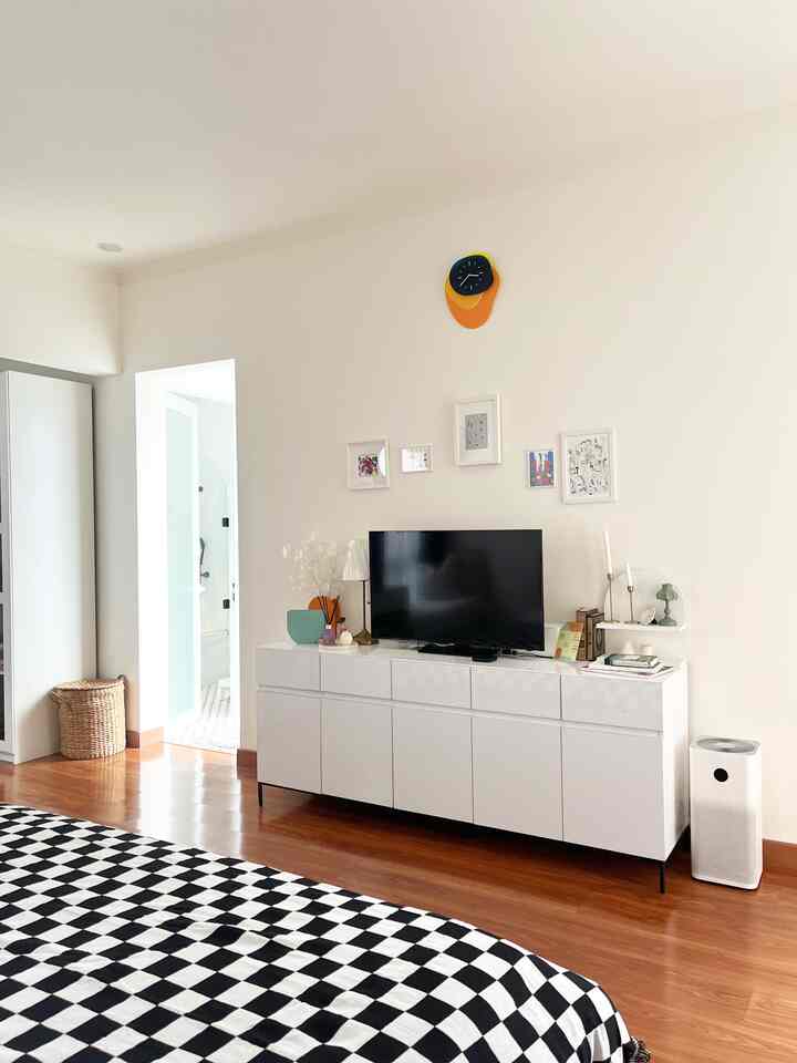 Bright white walls and light brown parquet floor bedroom featuring clean white dresser with TV, colorful wall clock and wall art in a minimal Scandinavian style space