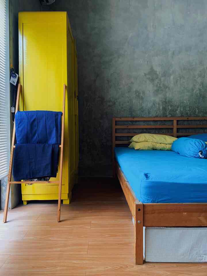 Wood tone bedroom featuring vibrant yellow wardrobe and blue bedding, with natural concrete walls and blinds creating a cozy atmosphere
