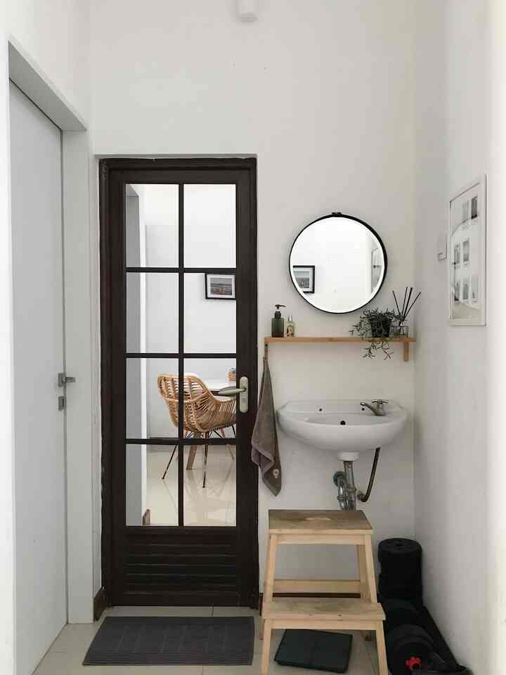 White and brown toned narrow bathroom with round mirror, wall-mounted sink, and wooden stool featuring a natural atmosphere