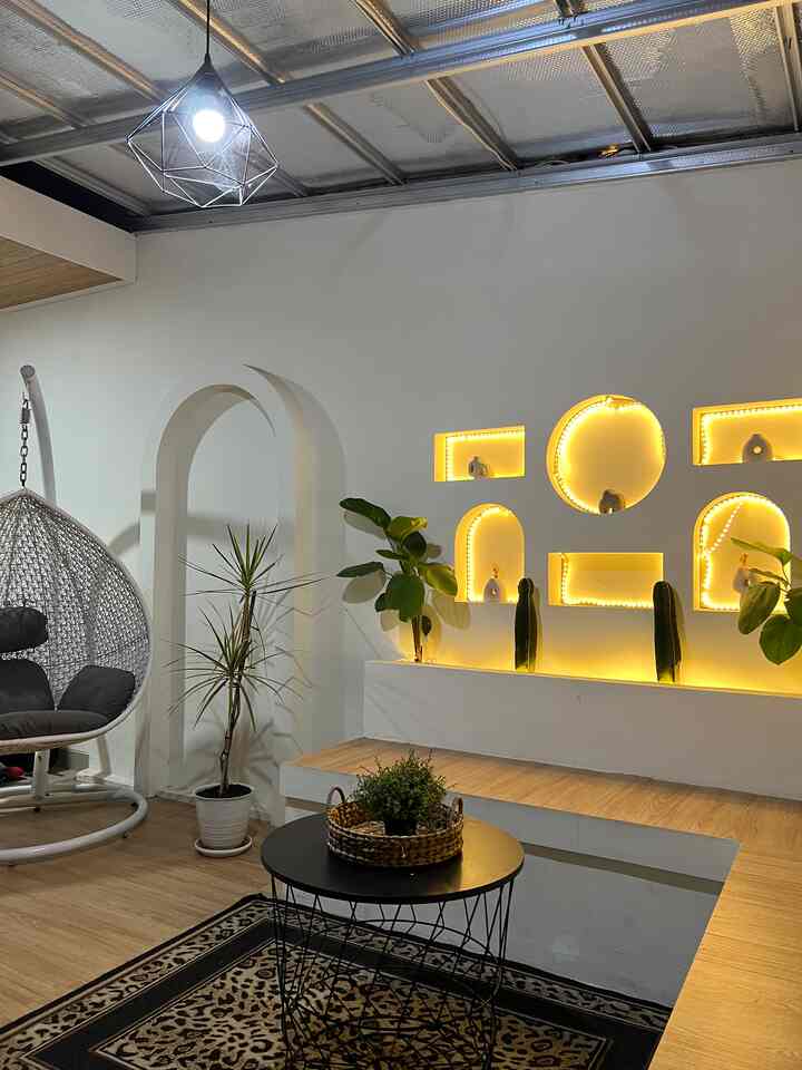 Warm natural-toned terrace featuring white walls, wooden floor, rattan hanging chair, plants, and ambient cafe-style lighting