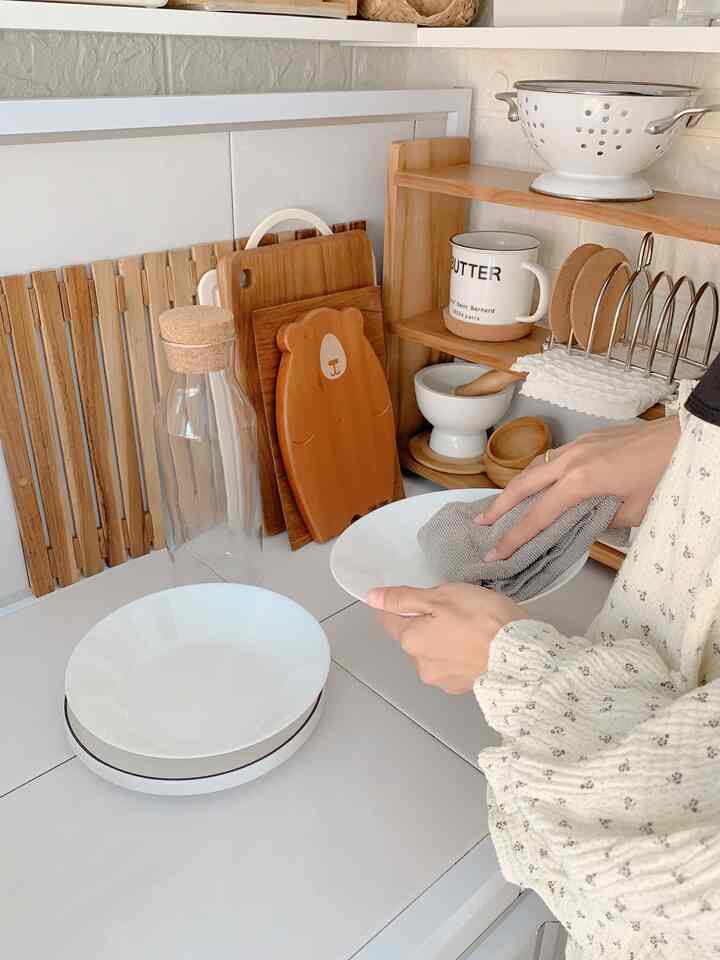 Bright white tile kitchen with wood tone cutting boards and shelves, hands cleaning a plate, cozy organized space