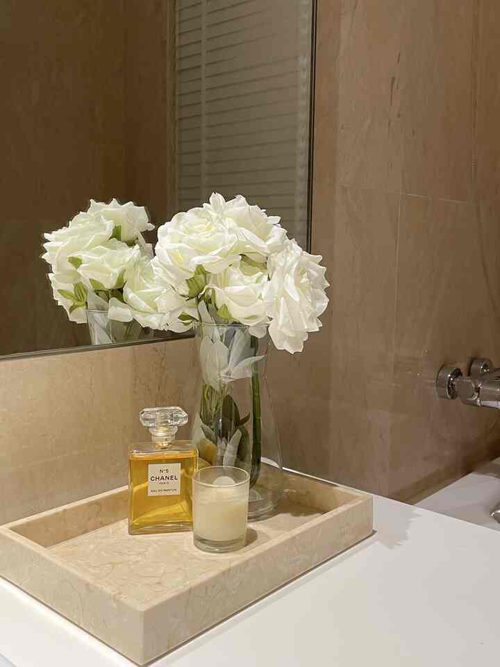 Beige tiled wall and white vanity topped with marble tray, white rose vase, perfume, and candle in a luxurious bathroom space