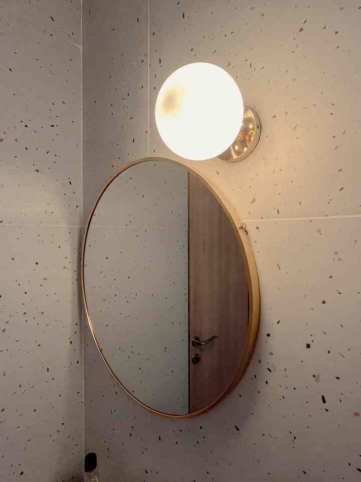 Minimal bathroom wall featuring terrazzo-patterned tiles, a gold-framed round mirror, and a spherical wall light