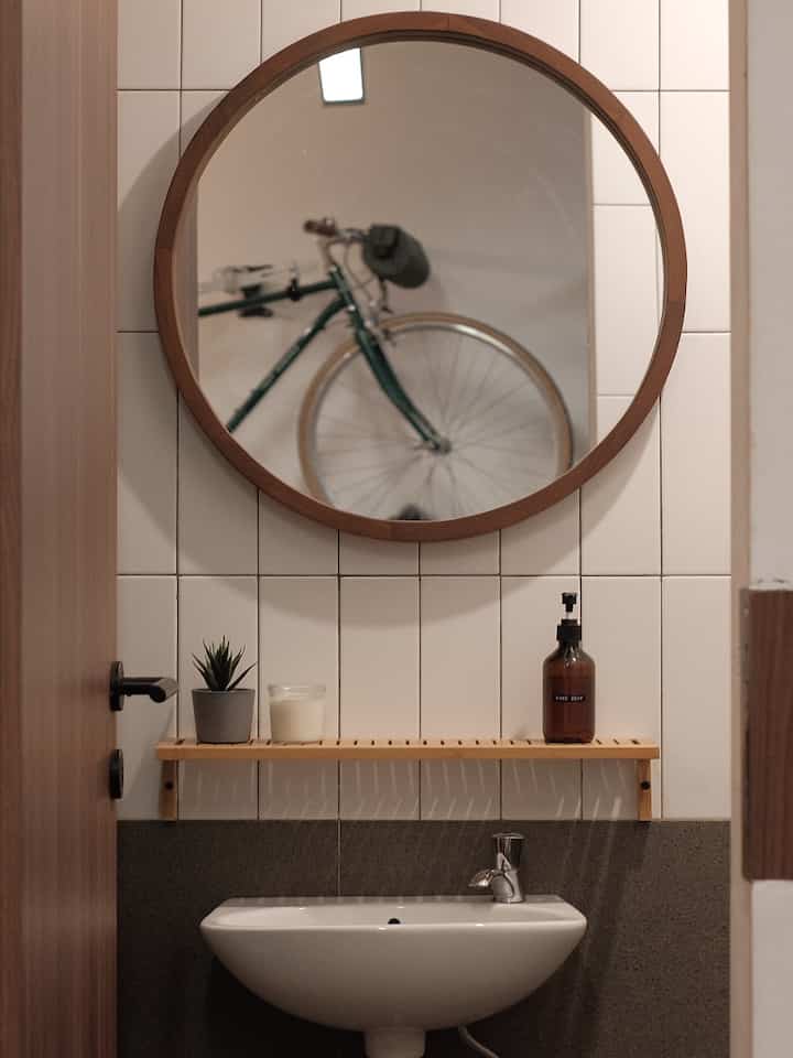 Natural wood tone round mirror and wooden shelf in a small white-tiled bathroom space