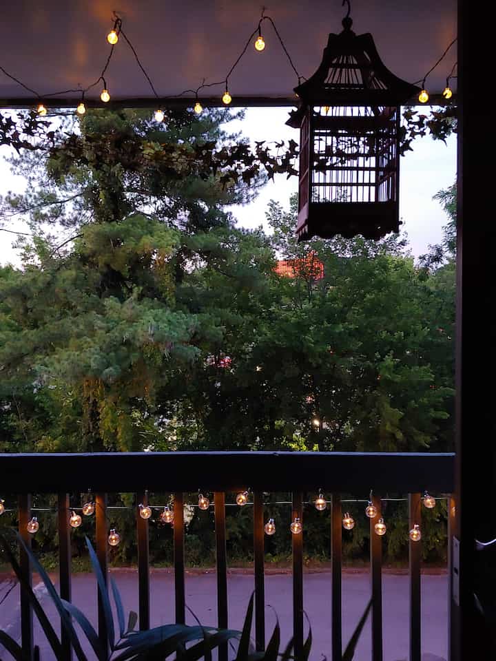 Evening balcony view with green trees visible outside, string lights hung on ceiling and railing creating a cozy ambiance