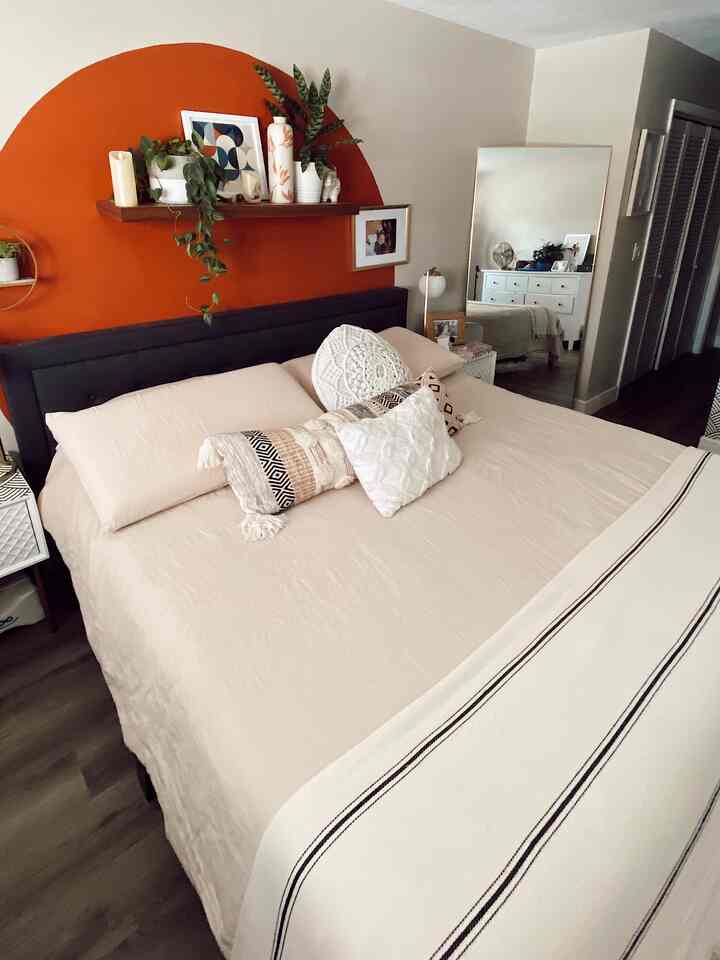 Natural style bedroom featuring beige bedding on dark bed frame, orange circular accent wall, floor lamp, and shelving with plants