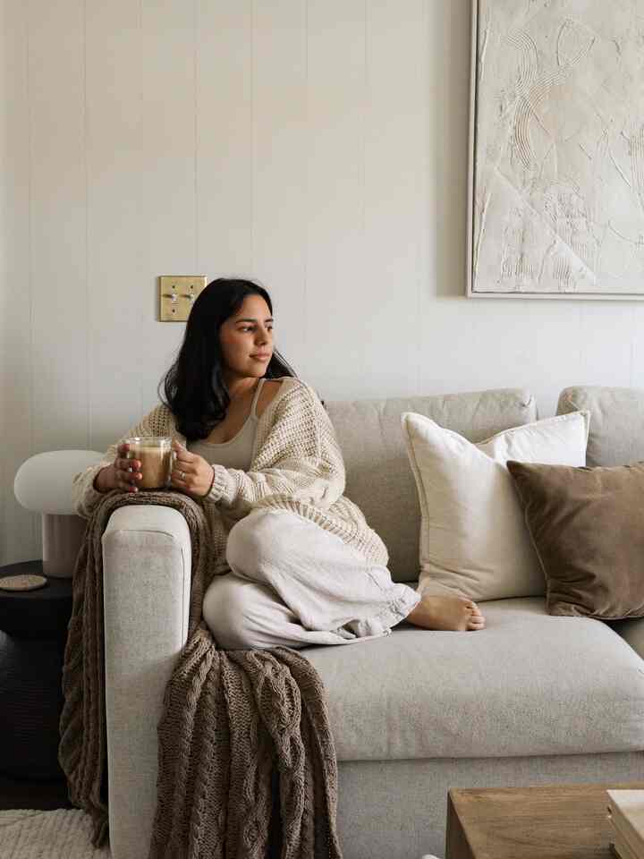 Natural beige and white toned living room featuring a sofa with cushions and a blanket where a woman relaxes sipping coffee in a cozy space